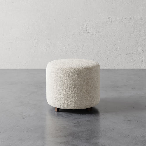 Lemieux et Cie Fabien 22" Round Ottoman in ivory bouclé, low cylindrical ottoman shown on concrete floor against textured plaster wall.