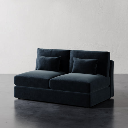 Lemieux et Cie Lacroix Armless Loveseat in deep teal, modular sofa with bolster pillows, shown on dark concrete floor against textured plaster walls.