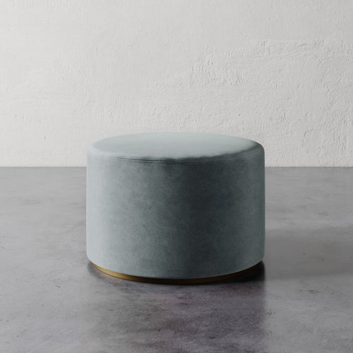 Lemieux et Cie Sophie Round Ottoman in sage upholstery with brass base detail, cylindrical ottoman on concrete floor against textured plaster wall.