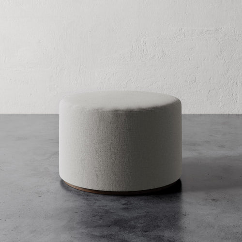 Lemieux et Cie Sophie Round Ottoman in neutral upholstery, low cylindrical form on stone floor against textured plaster wall.