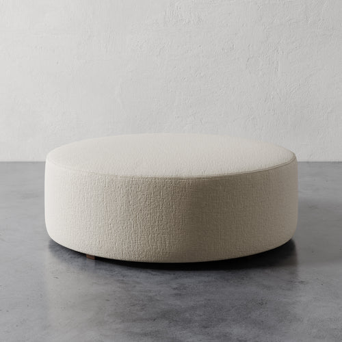 Lemieux et Cie Fabien 48" Round Ottoman in ivory upholstery, low-profile ottoman on polished concrete floor against textured plaster walls.