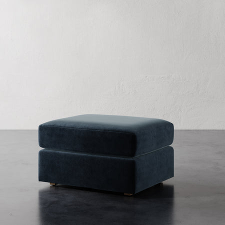 Lemieux et Cie Lacroix Ottoman in deep blue velvet, low-profile upholstered ottoman on dark concrete floor against textured plaster walls.