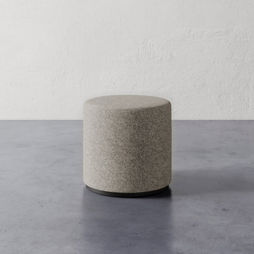 Lemieux et Cie Sophie Round Ottoman in neutral upholstery with dark base, cylindrical ottoman on concrete floor against textured plaster walls.
