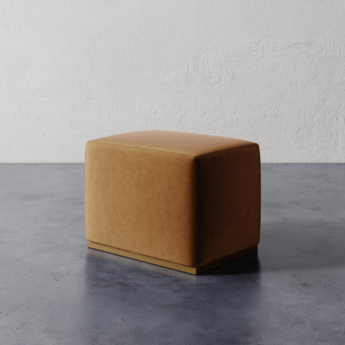 Lemieux et Cie Sophie Rectangular Ottoman in warm tan upholstery with slim base, compact seating piece on concrete floor against textured plaster walls.