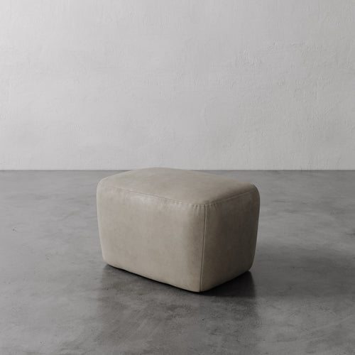 Lemieux et Cie Aurélie Leather Ottoman in warm neutral leather, softly rounded form on concrete floor against textured plaster wall.