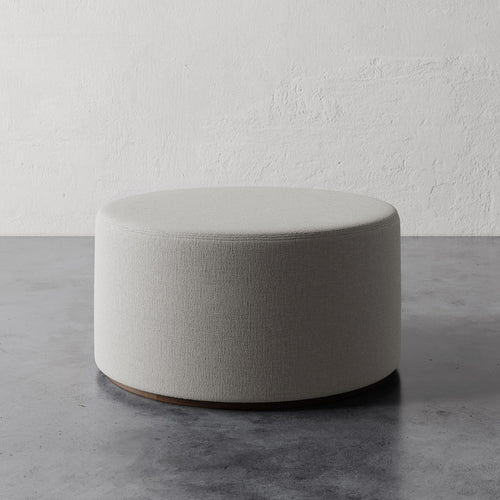 Lemieux et Cie Sophie Round 36" Ottoman in neutral upholstery, low rounded ottoman on concrete floor against textured plaster wall in soft natural light.
