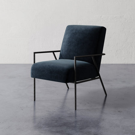 Lemieux et Cie Imane Lounge Chair in dark upholstery with slim dark frame, lounge chair on concrete floors against textured white plaster wall.