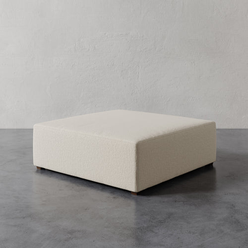 Lemieux et Cie Fabien 48" Square Ottoman in ivory, low-profile upholstered ottoman on polished concrete floor against textured plaster walls.