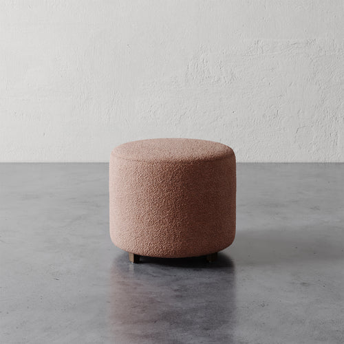 Lemieux et Cie Fabien 22" Round Ottoman in blush bouclé, cylindrical upholstered ottoman on dark polished concrete floor against textured plaster wall.