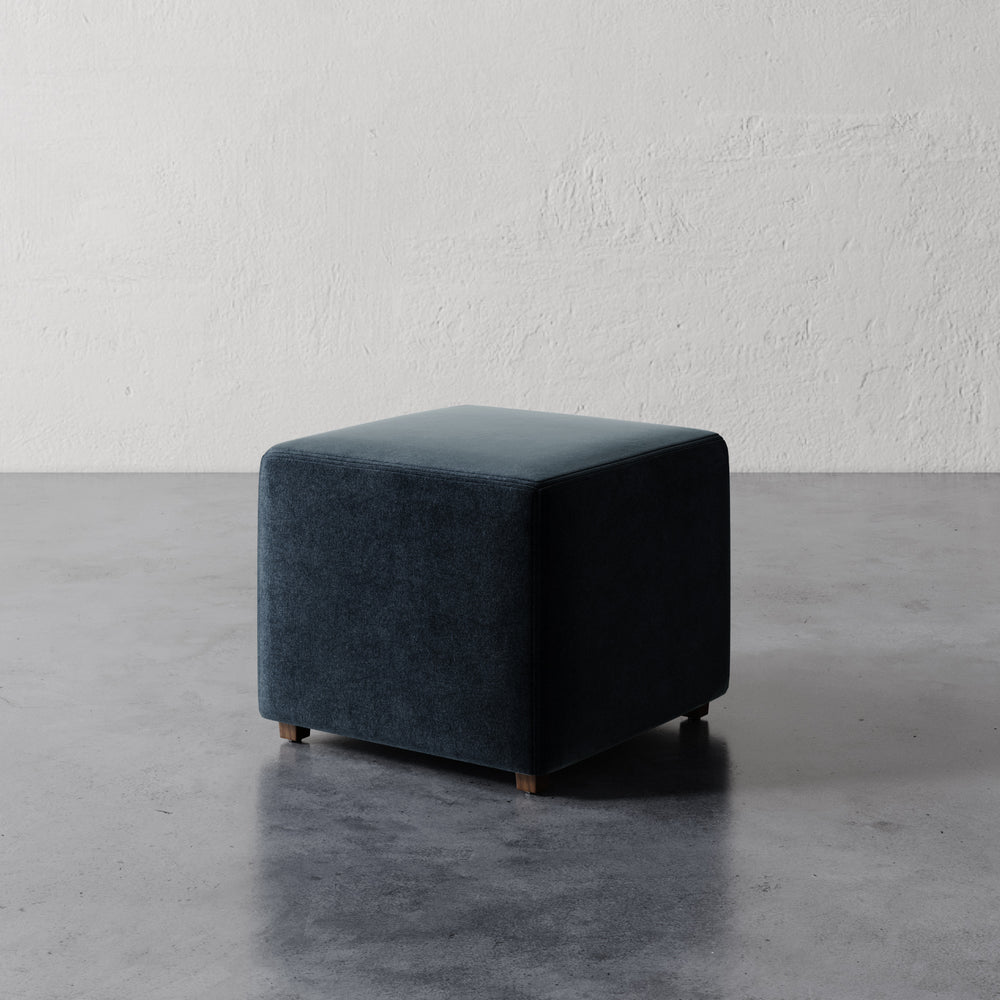 Lemieux et Cie Fabien 22" Square Ottoman in dark teal upholstery, low-profile seating on concrete floor against textured plaster walls.