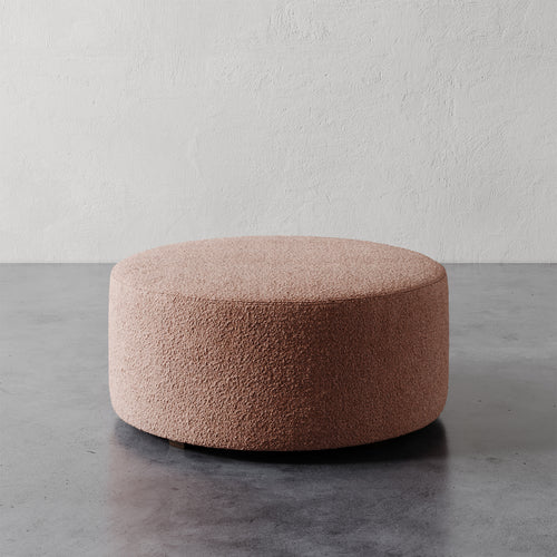 Lemieux et Cie Fabien 36" Round Ottoman in blush bouclé, low-profile round ottoman on concrete floor against textured plaster walls.