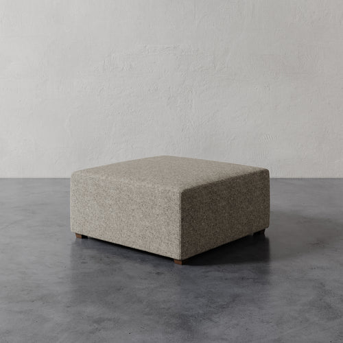 Lemieux et Cie Fabien 36" Square Ottoman in neutral upholstery, low-profile ottoman on dark concrete floor against textured plaster walls.