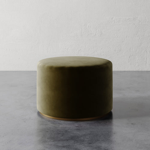 Lemieux et Cie Sophie Round Ottoman in olive upholstery with brass base detail, cylindrical ottoman on concrete floor against textured plaster wall.