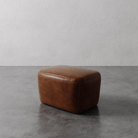 Lemieux et Cie Aurélie Leather Ottoman in warm cognac leather, low-profile ottoman with rounded edges on concrete floor against textured white plaster walls.