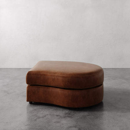 Lemieux et Cie Rulmann Right Ottoman in warm brown leather, low-profile curved ottoman on concrete floor against neutral walls.