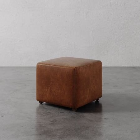 Lemieux et Cie Fabien Square Ottoman in warm brown leather, low-profile cube form on concrete floor against textured plaster wall.