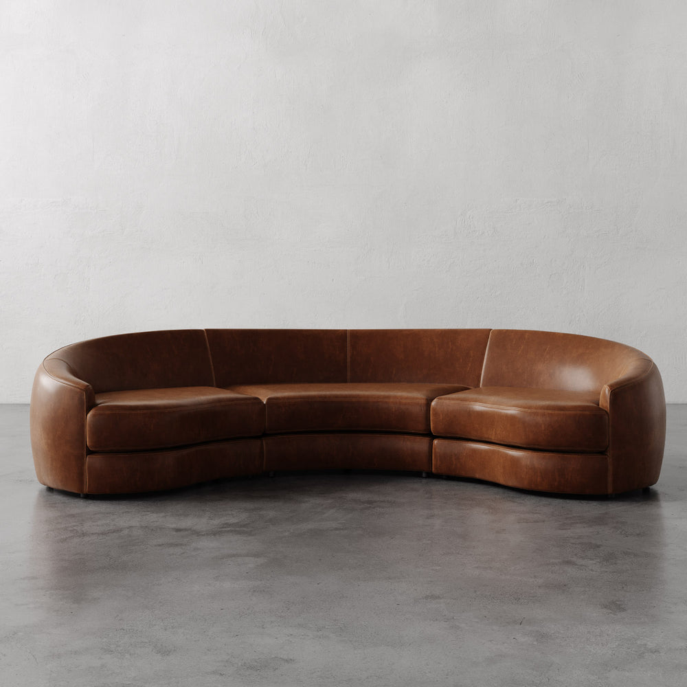 Lemieux et Cie Rulmann 3-Piece Sectional in warm tobacco, curved sofa with rounded arms on polished concrete floor and white plaster walls.