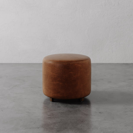 Lemieux et Cie Fabien Round Ottoman in warm tan leather, cylindrical ottoman with rounded top, on concrete floor against textured plaster wall.