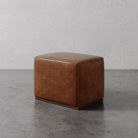 Lemieux et Cie Sophie Leather Rectangular Ottoman in warm tan leather, low-profile ottoman on concrete floor against textured plaster walls.