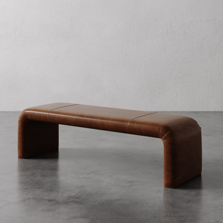 Lemieux et Cie Nadia Leather Bench in warm brown leather, low-profile bench with rounded sculptural form, shown on polished concrete floor against textured plaster walls.