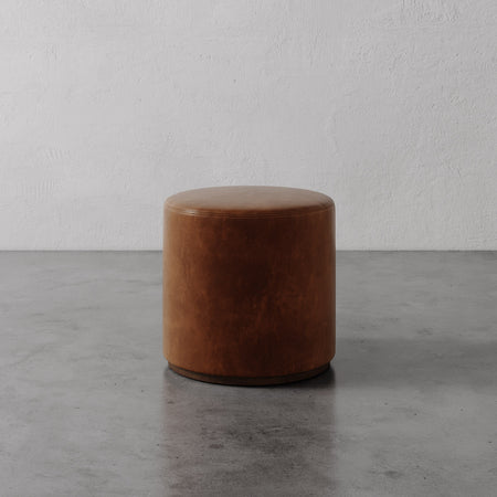 Lemieux et Cie Sophie Round Ottoman in tan leather, cylindrical ottoman with dark base, shown on concrete floor against textured plaster walls.