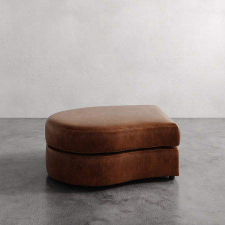 Lemieux et Cie Rulmann Left Ottoman in warm cognac leather, low-profile curved ottoman on concrete floor against neutral plaster walls.