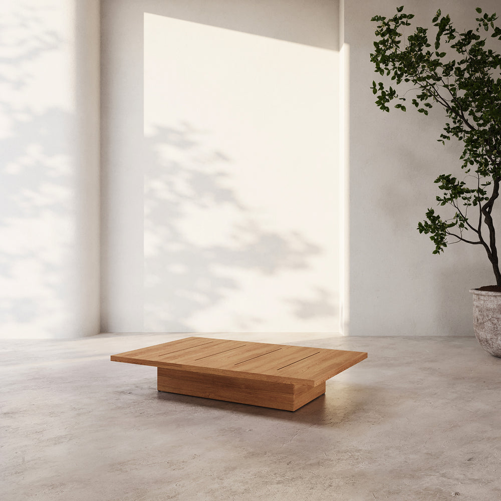 Lemieux et Cie S2 Teak Rectangular Coffee Table in solid teak, low-profile outdoor coffee table on stone floor in sunlit courtyard with white walls.