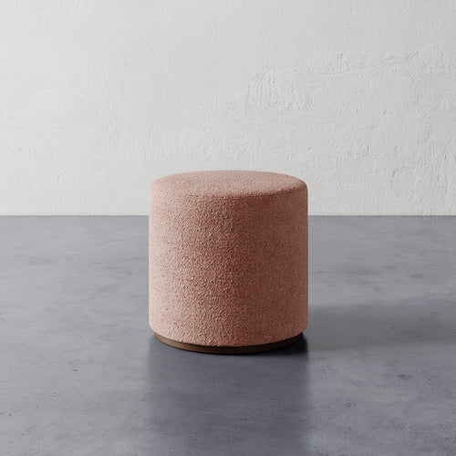 Lemieux et Cie Sophie Round Ottoman in blush upholstery, cylindrical ottoman on dark base, shown on concrete floor against textured plaster walls.