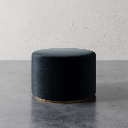 Lemieux et Cie Sophie Round Ottoman in dark teal upholstery with brass base, cylindrical ottoman on concrete floor against textured plaster wall.