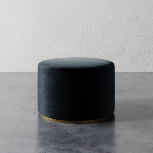 Lemieux et Cie Sophie Round Ottoman in dark teal upholstery with brass base, cylindrical ottoman on concrete floor against textured plaster wall.