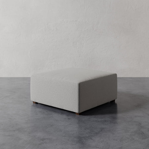 Lemieux et Cie Fabien 36" Square Ottoman in neutral upholstery, low-profile ottoman on wood legs, shown on concrete floor against textured plaster walls.