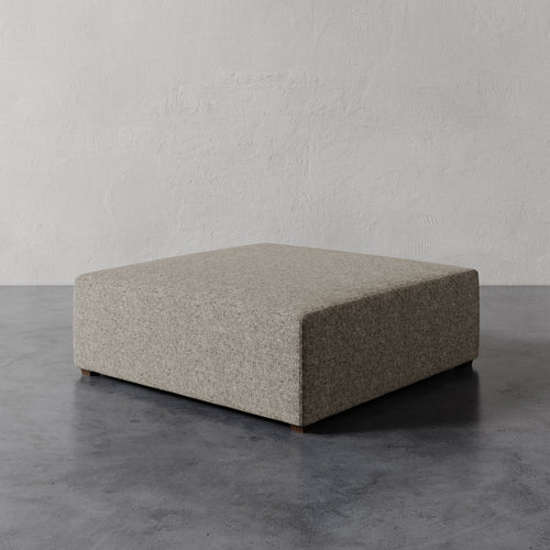 Lemieux et Cie Fabien 48" Square Ottoman in neutral taupe, low-profile upholstered ottoman on dark concrete floor against textured plaster walls.