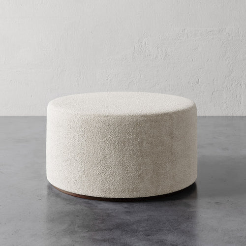 Lemieux et Cie Sophie Round 36" Ottoman in ivory textured upholstery, low-profile cylindrical ottoman on stone floor against textured plaster wall.