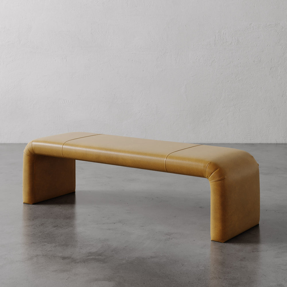 Lemieux et Cie Nadia Leather Bench in warm tobacco leather, low-profile bench with rounded sculptural form, shown on concrete floors against textured plaster walls.
