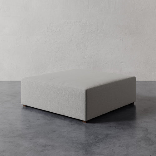 Lemieux et Cie Fabien 48" Square Ottoman in pale neutral, low-profile upholstered ottoman on stone floor against textured plaster walls.