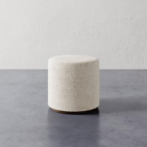 Lemieux et Cie Sophie Round Ottoman in ivory bouclé with dark wood base, cylindrical ottoman on concrete floor against textured plaster walls.