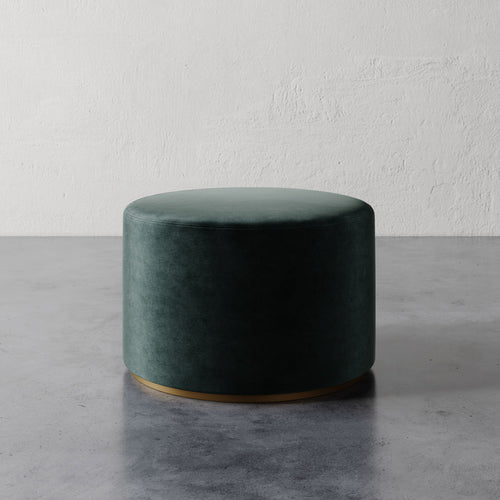 Lemieux et Cie Sophie Round Ottoman in deep green upholstery with brass base detail, cylindrical ottoman on polished concrete floor against textured plaster walls.