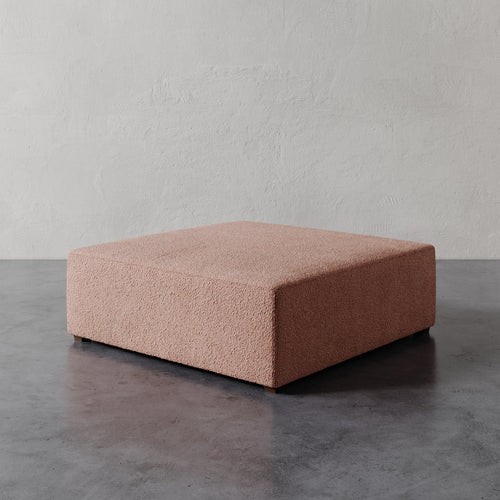 Lemieux et Cie Fabien 48" Square Ottoman in warm blush, low-profile geometric ottoman on dark concrete floor against textured plaster wall.