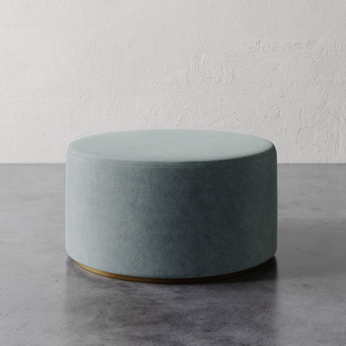 Lemieux et Cie Sophie Round 36" Ottoman in soft sage upholstery with brass base, shown on polished concrete floor against textured plaster walls.