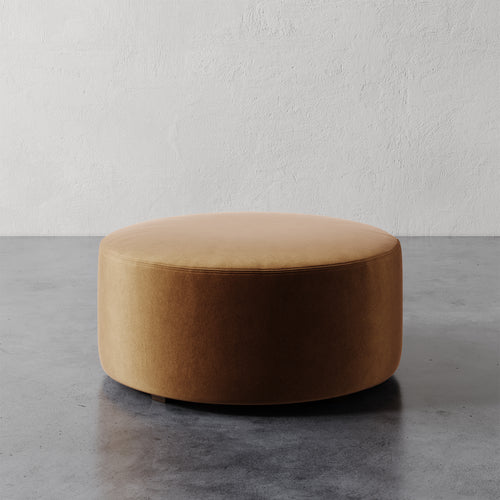 Lemieux et Cie Fabien 36" Round Ottoman in warm tan upholstery, round ottoman on gray concrete floor against textured white plaster wall.