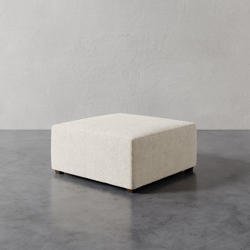 Lemieux et Cie Fabien 36" Square Ottoman in ivory textured upholstery, low-profile ottoman on stone floor against neutral plaster wall.