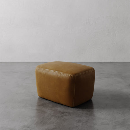 Lemieux et Cie Aurélie Leather Ottoman in warm camel leather, low-profile ottoman with rounded form, shown on concrete floor against textured plaster walls.
