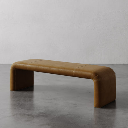 Lemieux et Cie Nadia Leather Bench in warm tan leather, low-profile bench with rounded waterfall edges, shown on polished concrete floor against textured plaster walls.