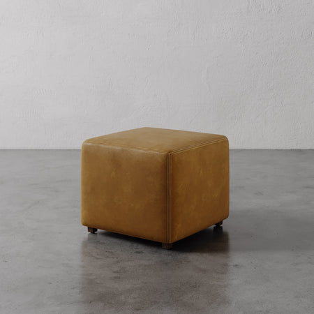 Lemieux et Cie Fabien Square Ottoman in warm cognac leather, low-profile cube form on stone floor against textured white walls.