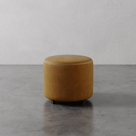 Lemieux et Cie Fabien Round Ottoman in warm caramel leather, cylindrical form on low legs, shown on concrete floor against textured plaster walls.