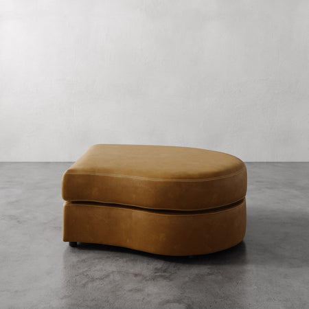 Lemieux et Cie Rulmann Right Ottoman in warm cognac leather, curved low-profile ottoman on stone floor against neutral plaster wall.
