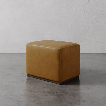 Lemieux et Cie Sophie Leather Rectangular Ottoman in warm tan leather with dark wood base, shown on concrete floor against textured plaster walls.