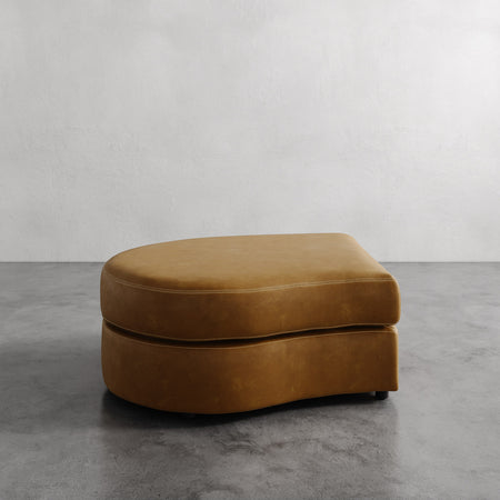 Lemieux et Cie Rulmann Left Ottoman in warm ochre, low-profile curved ottoman on concrete floor against neutral textured wall.