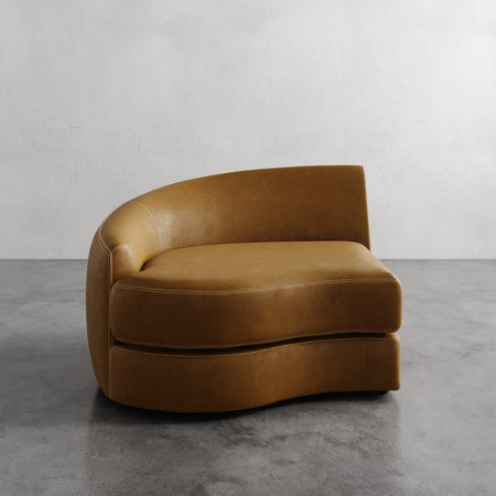 Lemieux et Cie Rulmann Left Arm Chair in warm caramel leather, curved sculptural form on concrete floor against white textured wall.