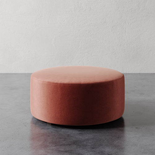 Lemieux et Cie Fabien 36" Round Ottoman in warm coral upholstery, low-profile seating piece on polished concrete floor against textured plaster wall.